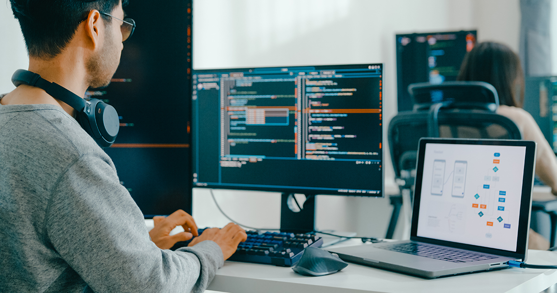Person typing on a keyboard in front of a monitor displaying code in a dark-themed editor.