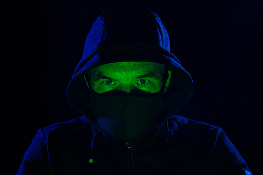 Man in a hooded jacket and mask lit dramatically in green and purple.