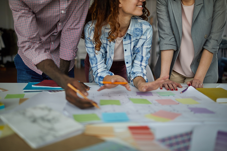 Close-up of hands and arms as a team works on a table covered with sticky notes, plans and diagrams.
