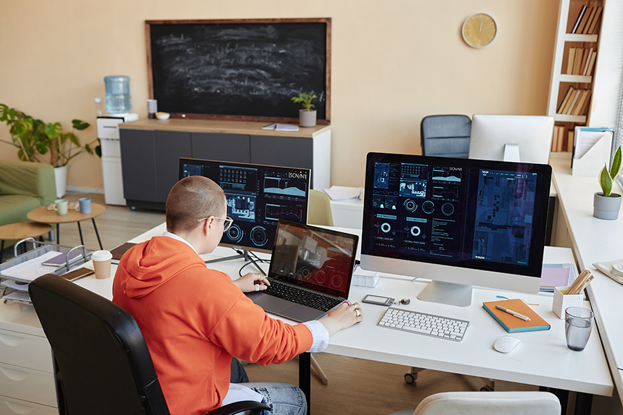 Developer in a red hoodie analysing data dashboards on multiple monitors.