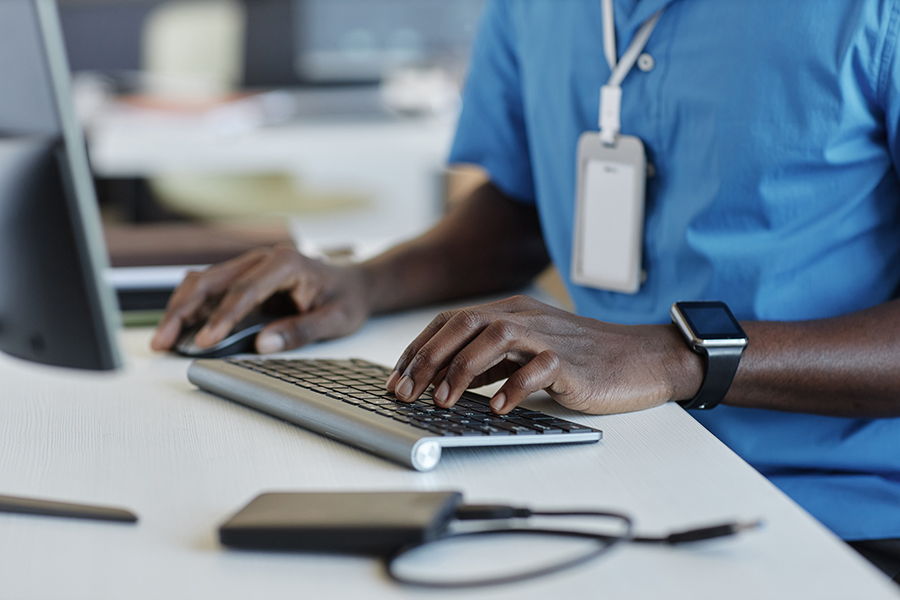 Close-up of hands typing on wireless keyboard, smartwatch and ID badge visible, in professional environment.