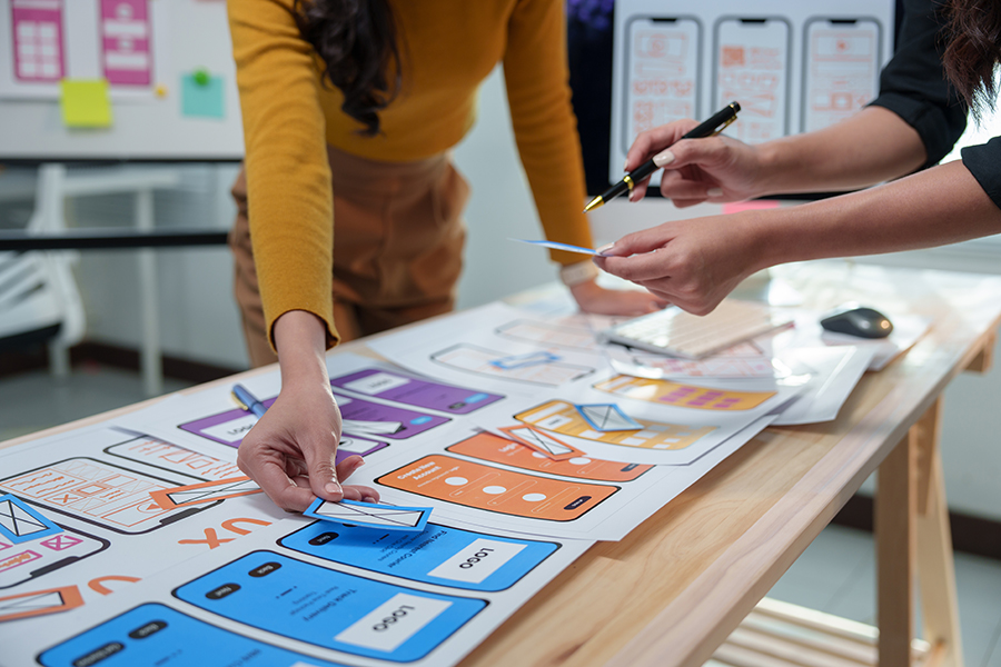 Hands arranging mobile app wireframes on a desk covered with UX layouts