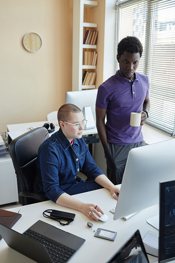 Two colleagues reviewing content on a desktop monitor, one seated, the other standing.