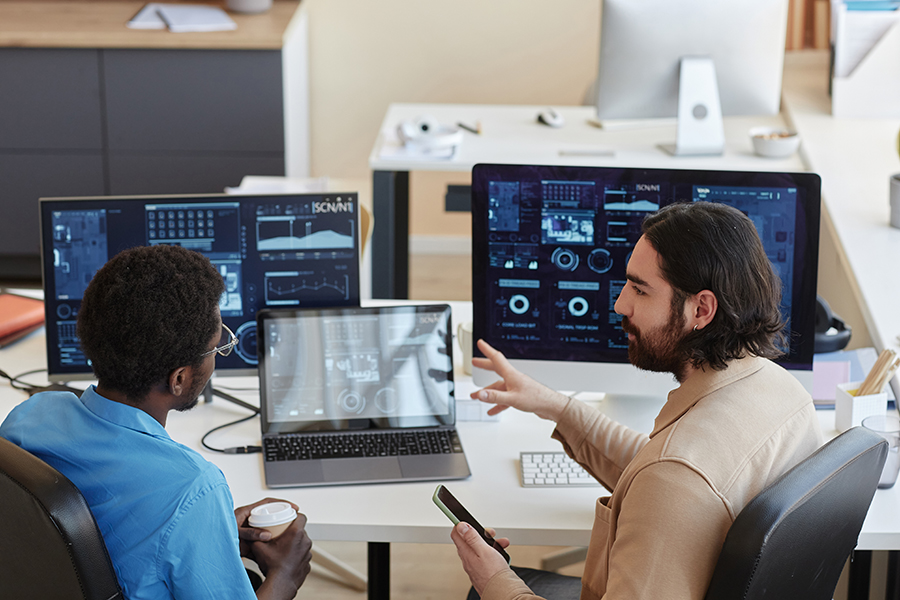 Two team members in conversation at a workstation, reviewing performance dashboards and charts on multiple screens.